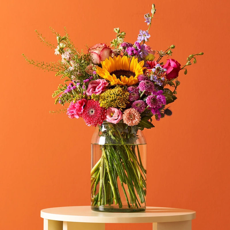 Boeket Altijd Zonnig met zonnebloem, allium en kleurrijke zomerbloemen op tafel voor oranje achtergrond