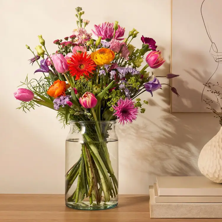 Kleurrijk seizoensboeket met gerbera, ranonkel en tulpen in glazen vaas op plank