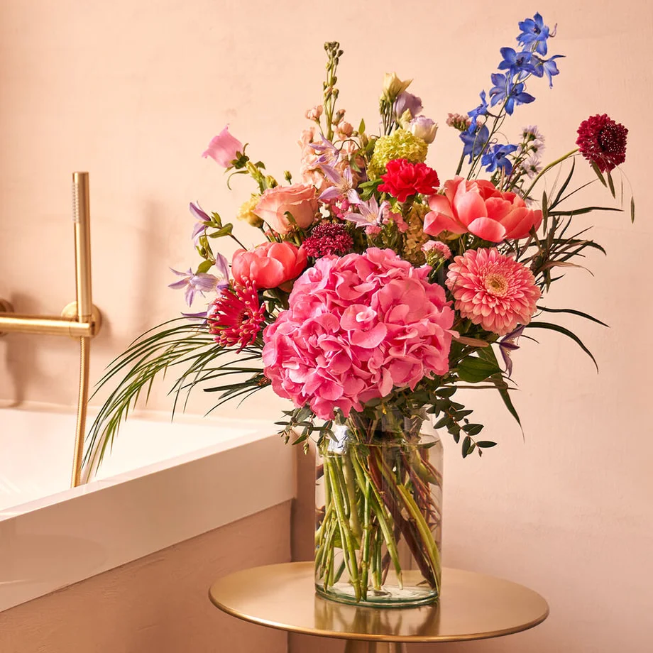 Boeket met roze hortensia, gerbera, rozen en blauwe delphinium in een glazen vaas op tafel in keuken