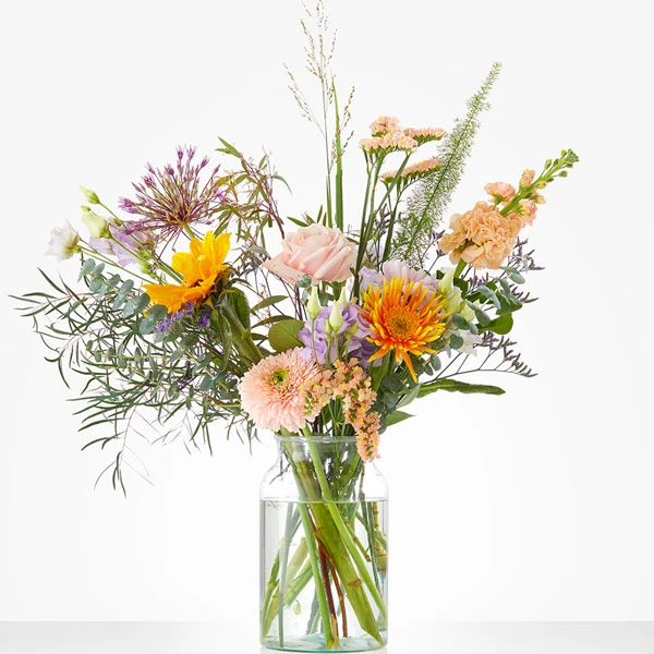 Speels voorjaarsboeket met rozen, gerbera’s, allium en diverse seizoensbloemen in warme en frisse tinten in glazen vaas
