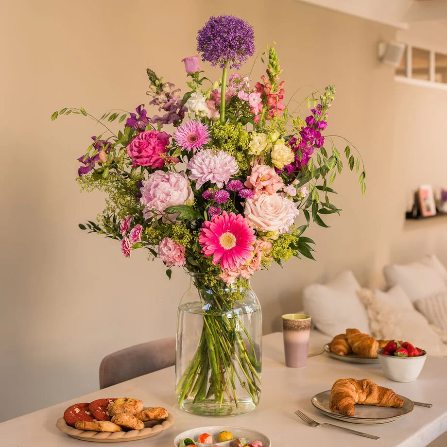 Boeket met rozen, gerbera, pioenroos, allium en leeuwenbek in een glazen vaas op feestelijk gedekte ontbijttafel
